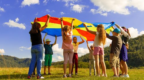 Group of kids playing with a rainbow parachute cloth in a field